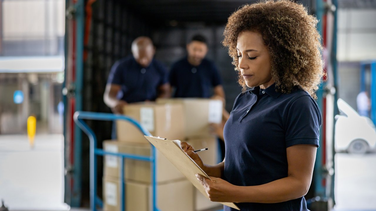 A woman with curly hair holds a clipboard in a shipping container, while two men unload boxes behind her.