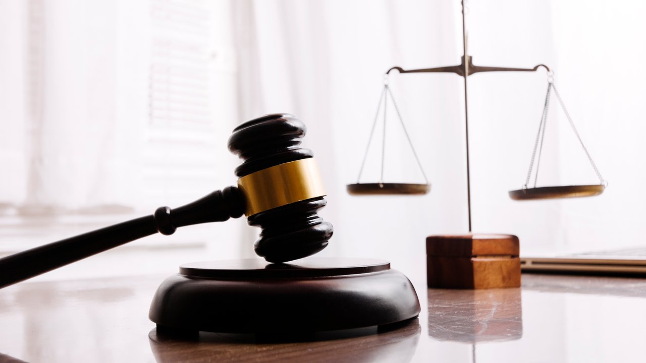 A wooden gavel and a balance scale on a reflective surface with soft lighting in the background.