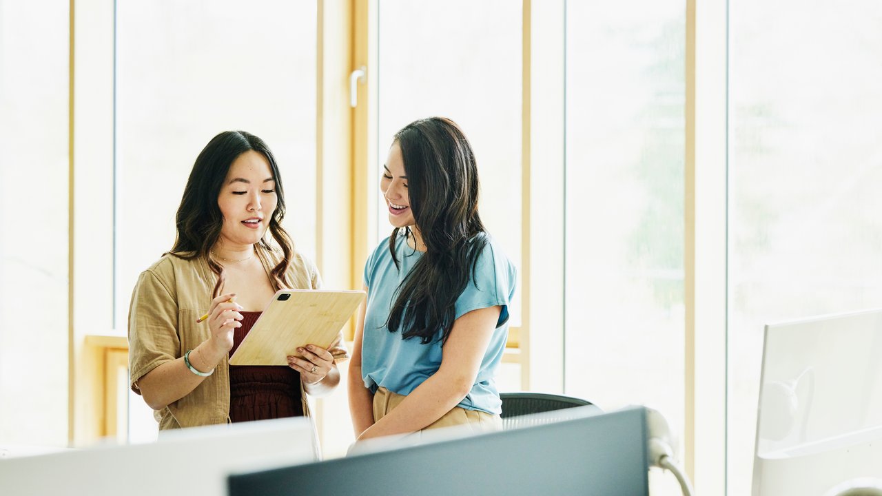 Two businesswomen working on digital tablet in open office sharing what they have learned about IDI and Group Disability