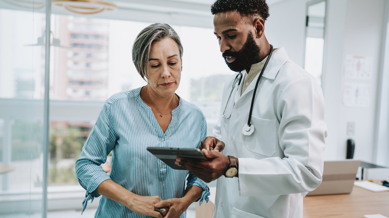 A doctor in a white coat discusses a tablet with a woman in a striped shirt in a medical office.