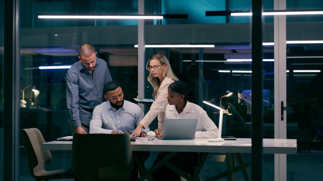 Four professionals collaborate at a table in a modern office, with laptops and documents in a well-lit environment.