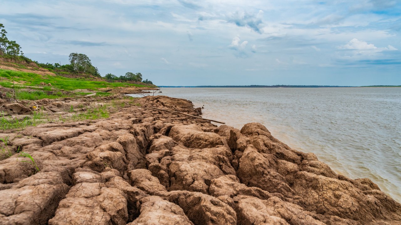 A riverbank with cracked earth and sparse vegetation under a cloudy sky.
