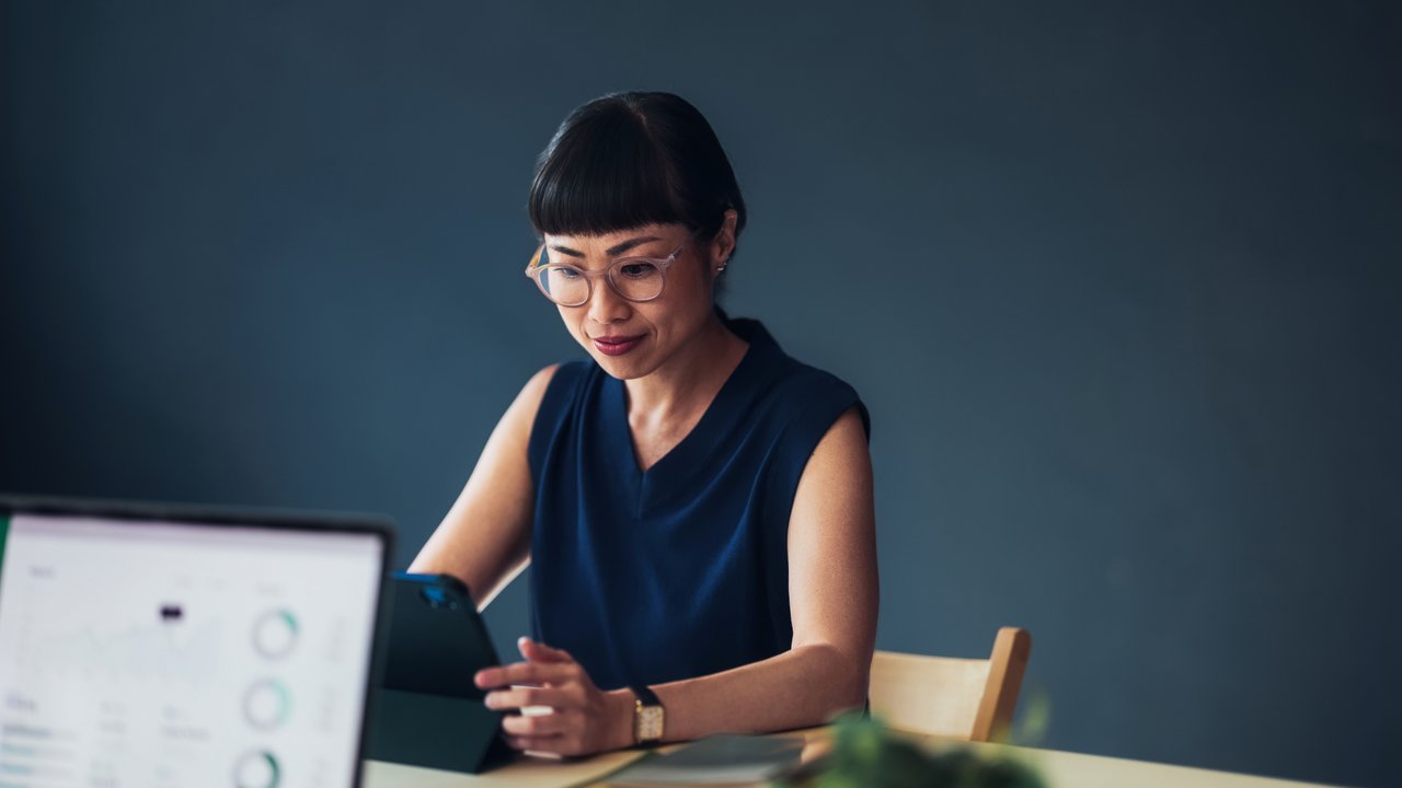 A woman in a navy top sits at a table, using a tablet, with a laptop and plants visible in the foreground.