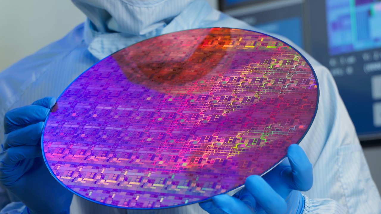 Asian male technician in sterile coverall holds wafer that reflects many different colors with gloves and check it at semiconductor manufacturing plant