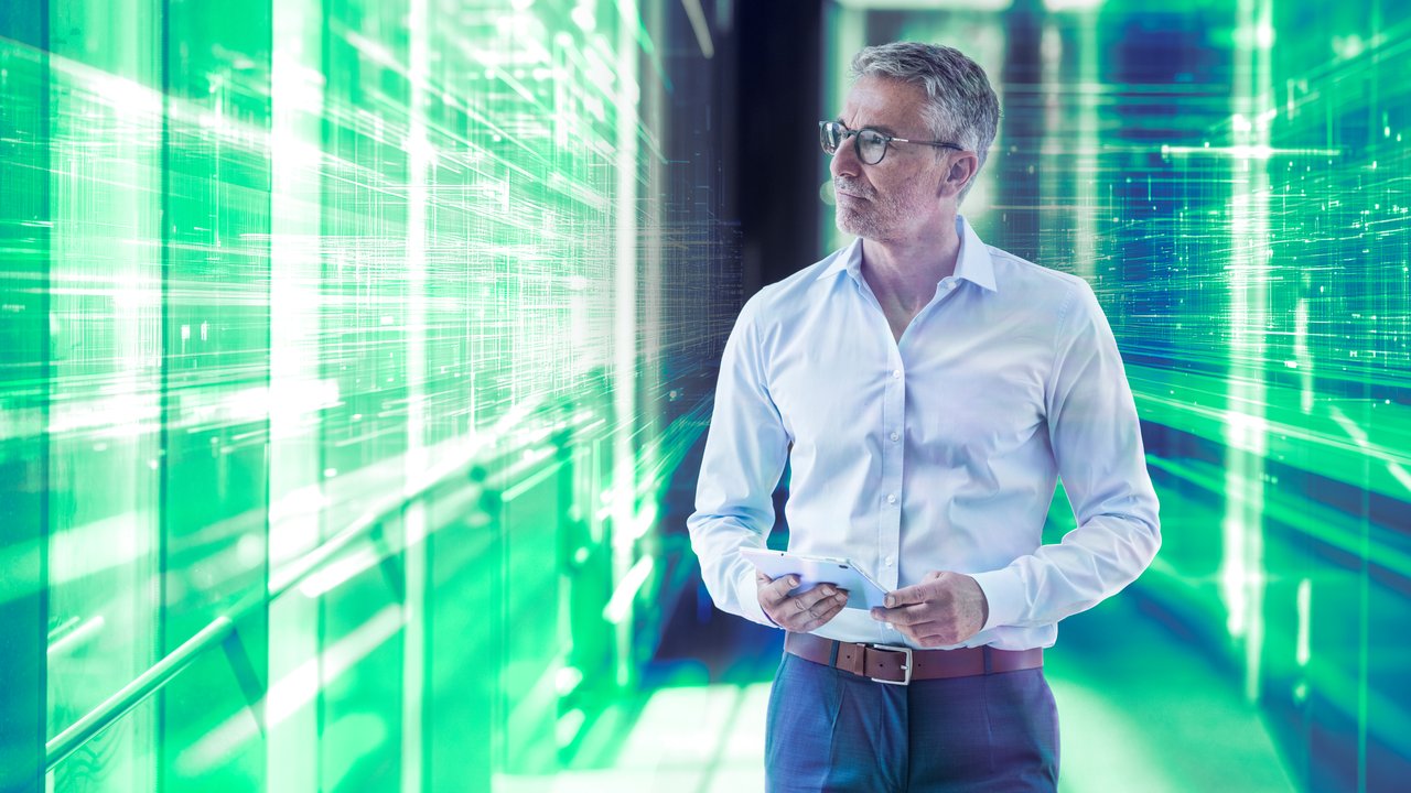 A man in a white shirt stands in a futuristic hallway with green digital patterns and holds a device.