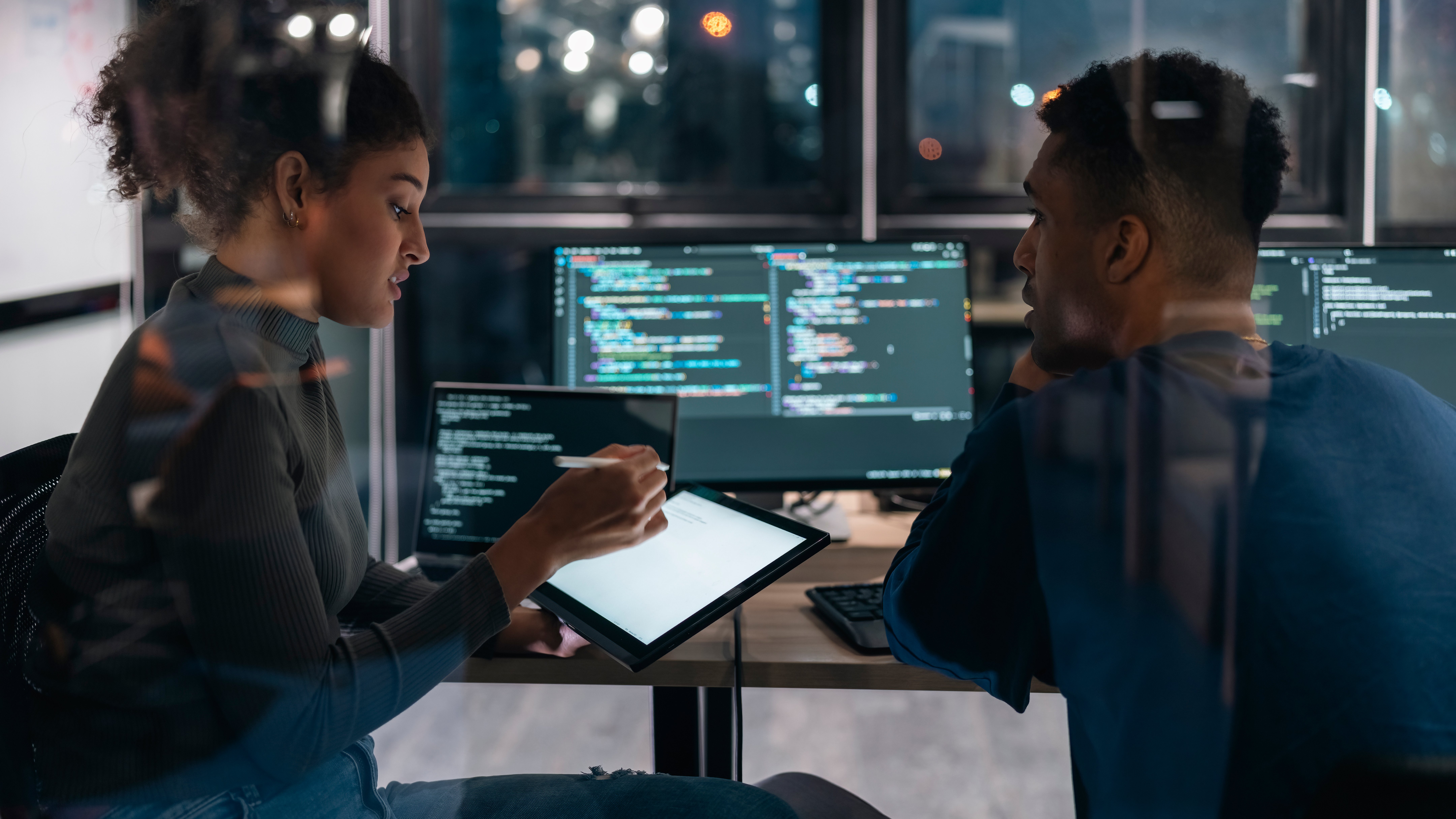 A woman discusses coding on a tablet with a man at a desk with two computer monitors displaying code.