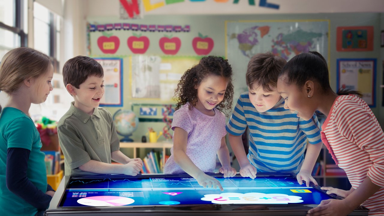 Five children interact with a touchscreen display in a classroom setting, with educational materials in the background.