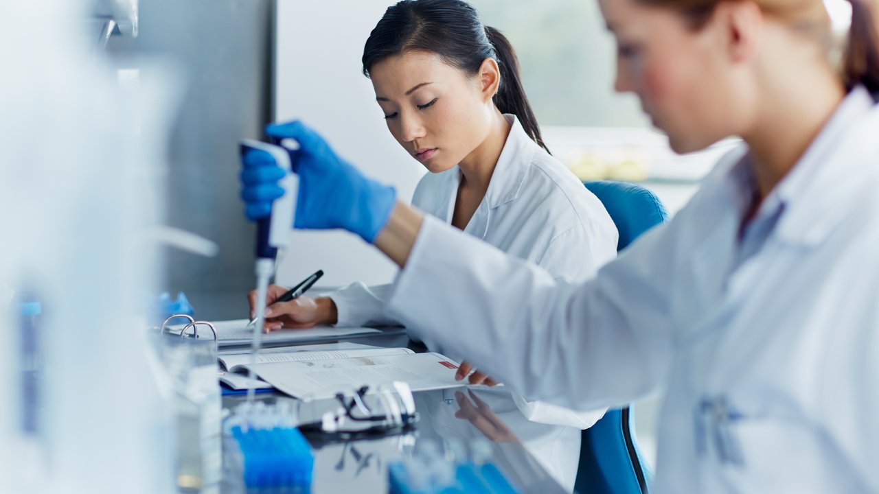 Two women in lab coats working in a laboratory, one using a pipette and the other writing notes for biological age testing.