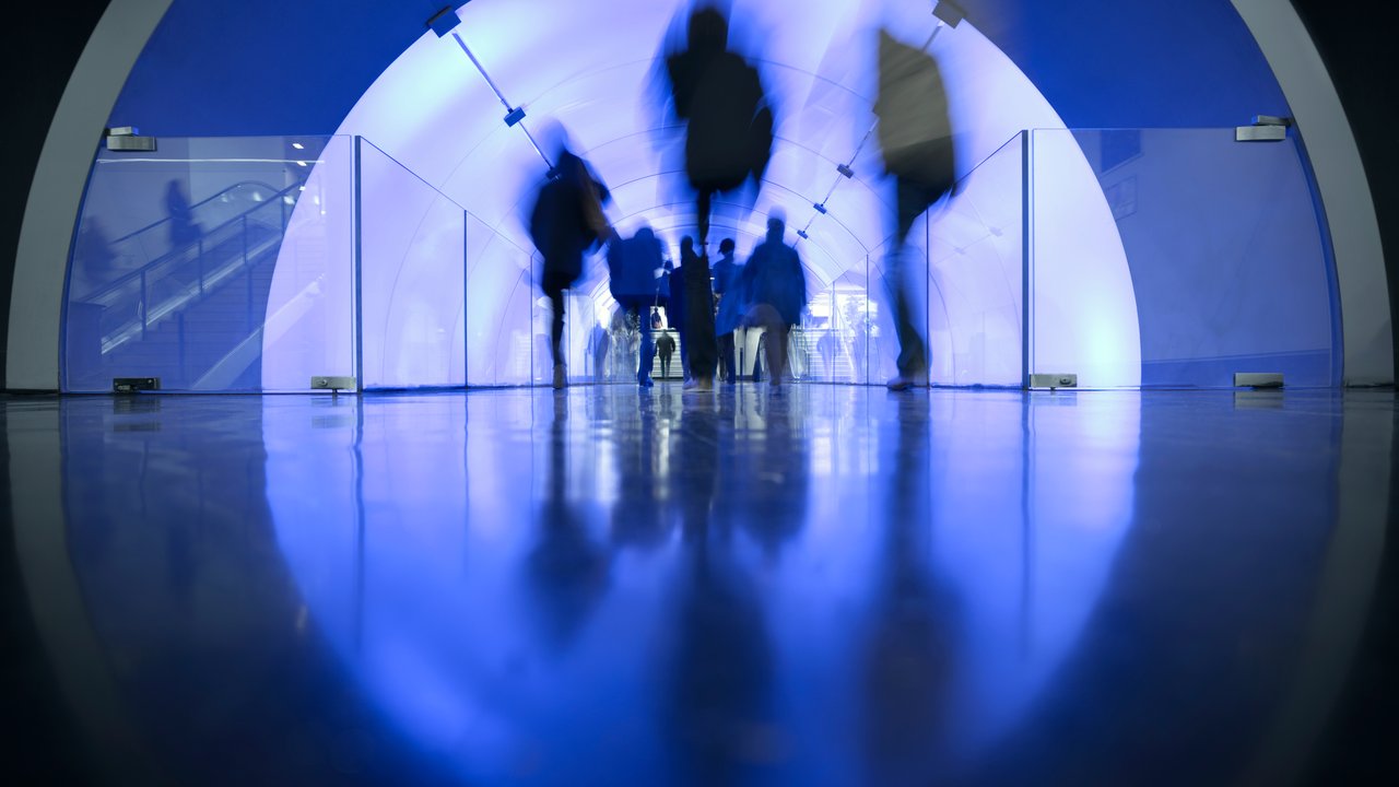 A blurred view of people walking through a blue-lit tunnel with glass barriers and a shiny floor.