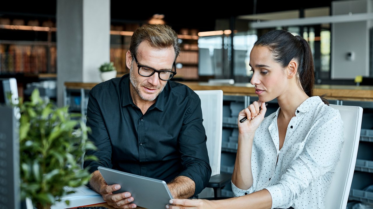 Colleagues Collaborating on a Tablet in Office A man and a woman sit at a table, looking at a tablet in an office setting.