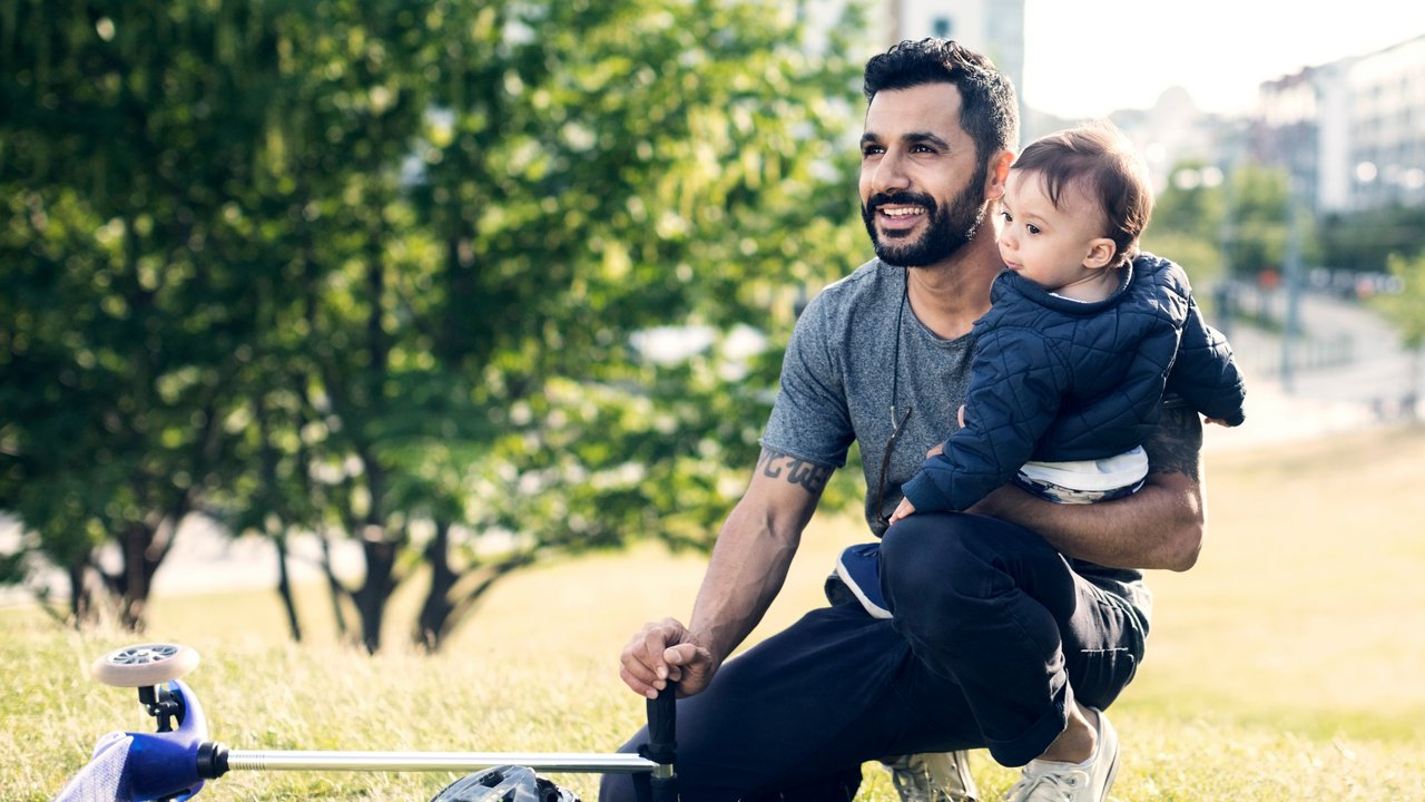 Father carrying toddler while sitting by push scooter on grassy field at park