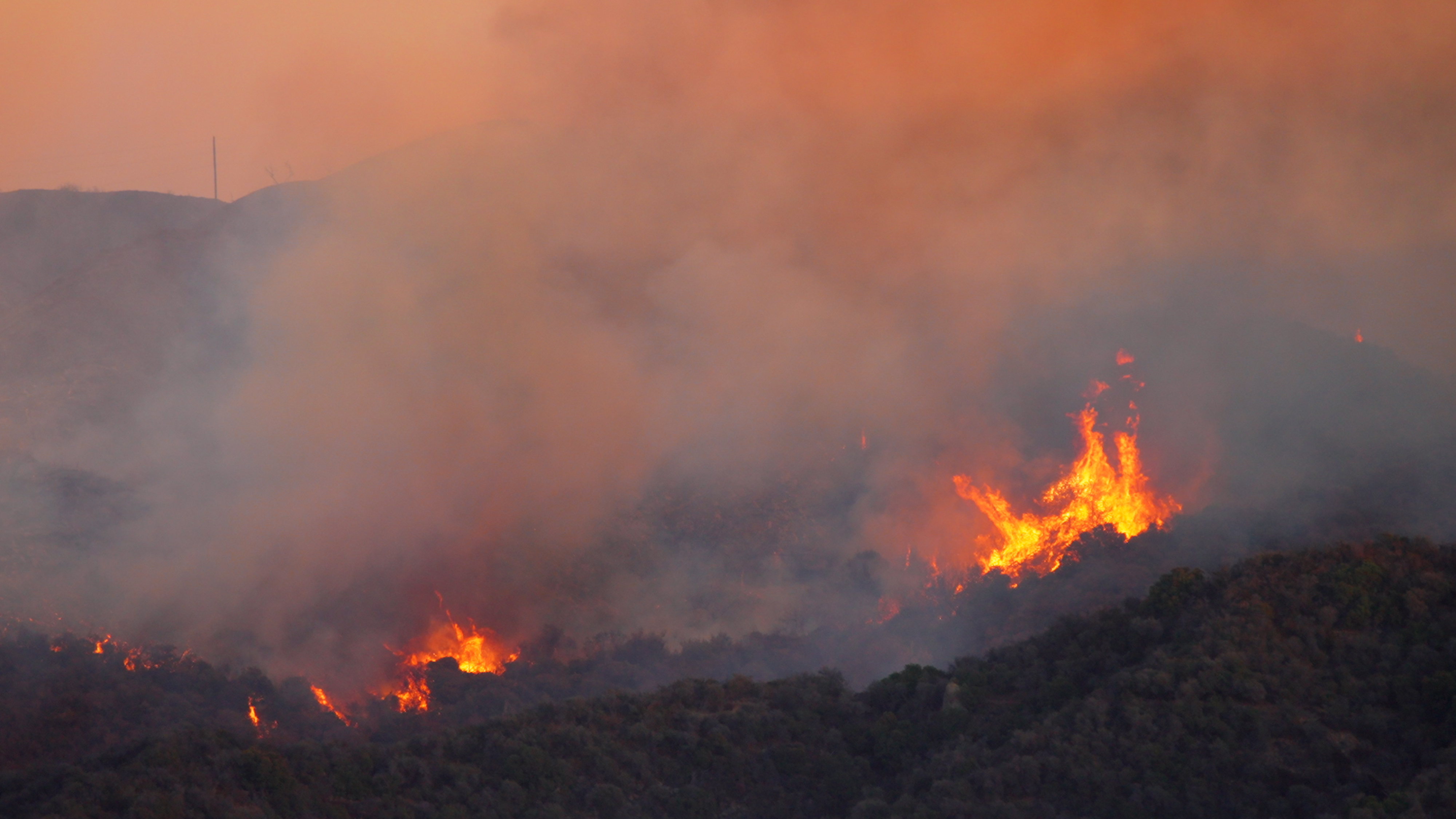 A wildfire burns on a hillside, surrounded by smoke and an orange sky at sunset.