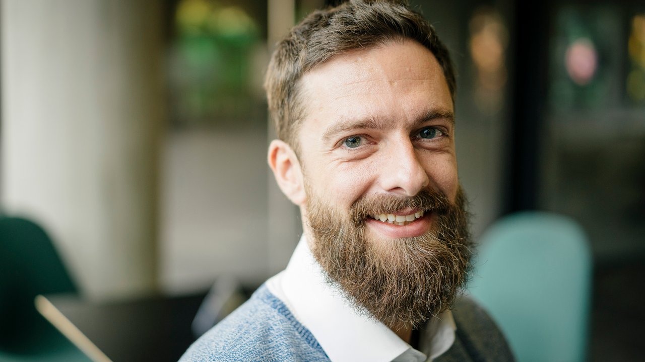 A smiling man with a beard wearing a blue sweater and white shirt, seated indoors with blurred background.