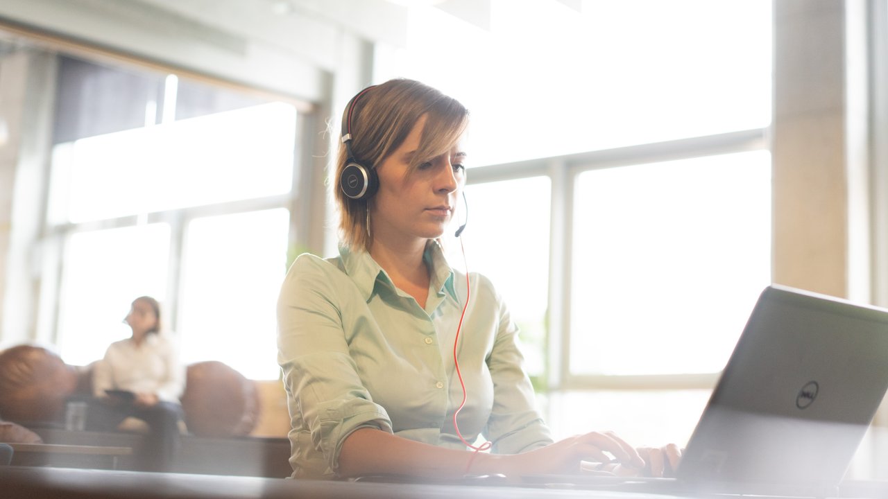 A woman in a light green shirt uses a laptop while wearing headphones in a bright, modern space.