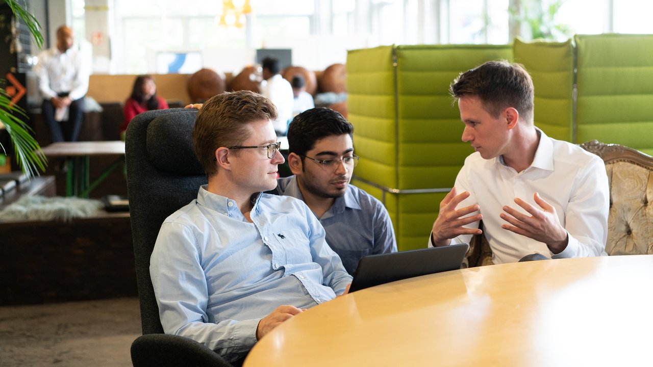 Three men discuss while seated at a round table with a laptop, in a modern office space.