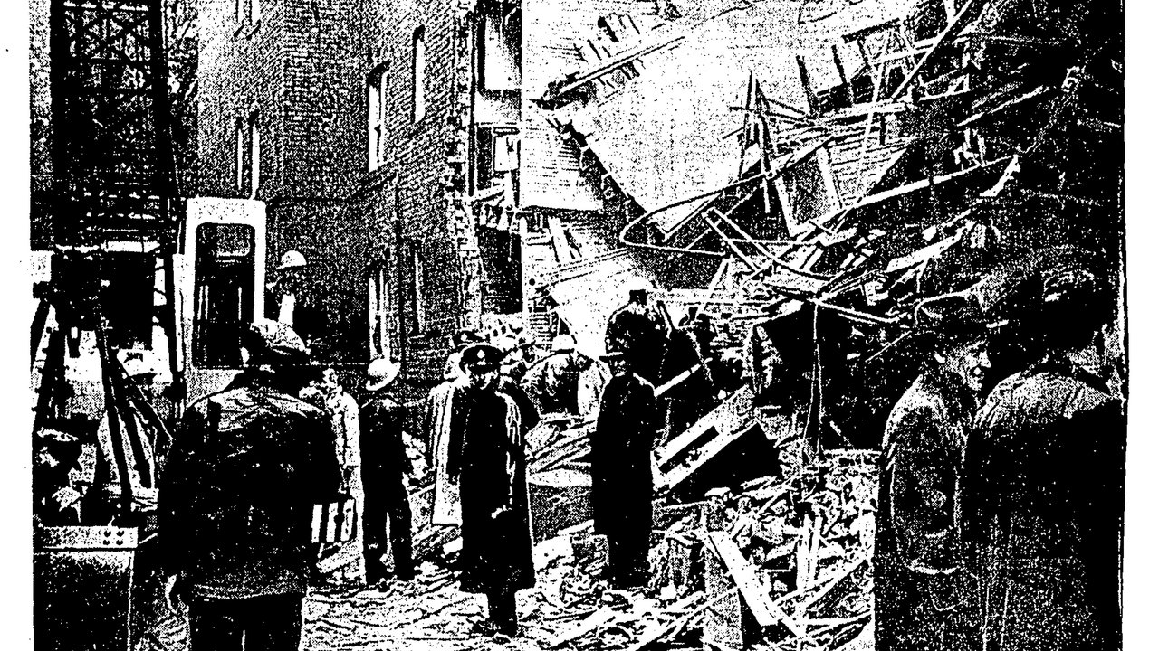 Amidst rubble and debris, a group of individuals gathers near a collapsed building. The scene is marked by a partially destroyed brick structure, with wooden beams and twisted metal scattered across the ground. Some people wear hard hats, indicating a response to an emergency. The atmosphere conveys urgency and concern.