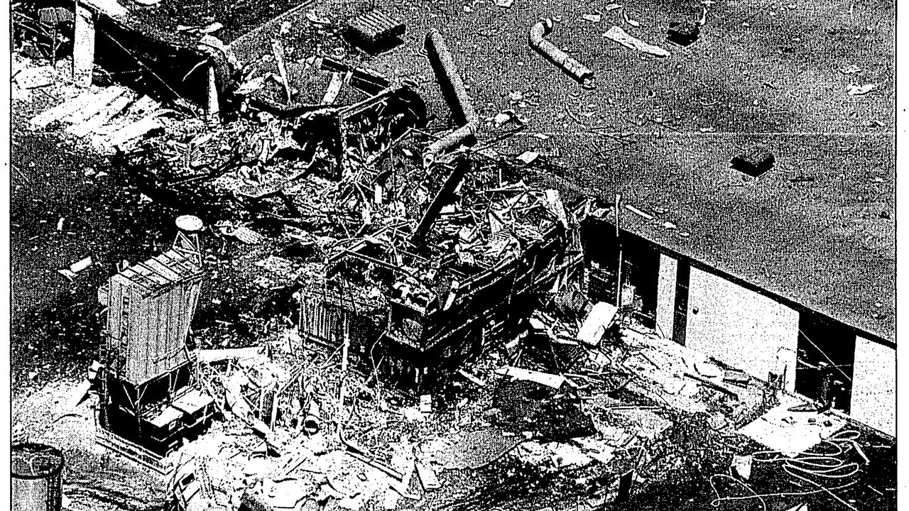 Aerial view of a devastated industrial site, with debris scattered across the ground. A large structure is partially collapsed, surrounded by twisted metal and rubble. Pipes and building materials are strewn about, indicating a recent explosion. The scene conveys destruction and urgency, highlighting the aftermath of a significant incident.