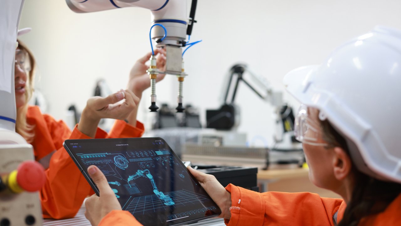 In a modern workspace, a robotic arm hovers above a table, equipped with a gripper. Two individuals in orange work attire interact with the robot, one holding a tablet displaying a digital interface of robotic controls. The background features additional robotic arms, emphasizing a high-tech environment.