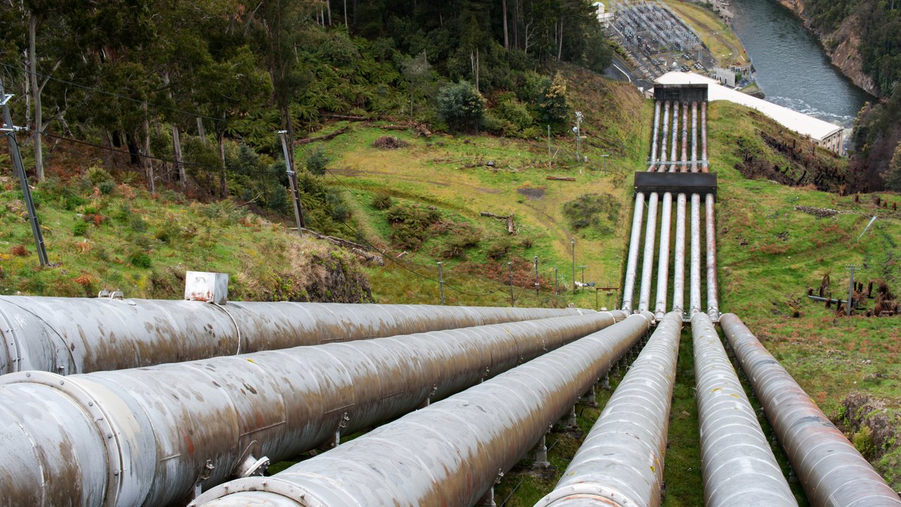 A series of large, metallic pipes extend down a hillside, leading towards a river and a power station in the distance. The landscape features lush greenery and scattered trees, with a gentle slope and some rocky areas. Power lines run alongside the pipes, contributing to the industrial setting amidst natural surroundings.