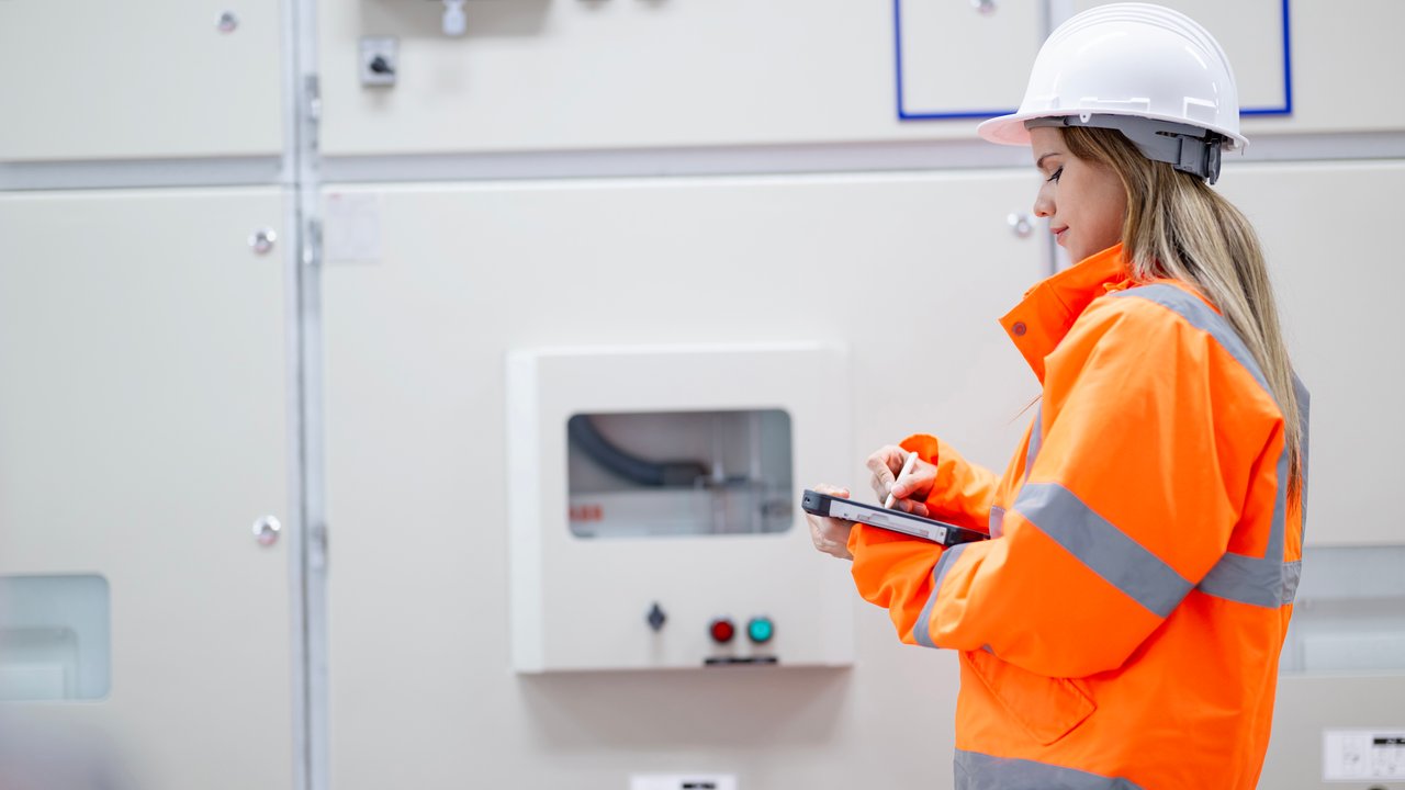 A person in a bright orange safety jacket with reflective stripes and a white hard hat stands in front of a large electrical panel. They hold a tablet and a pen, appearing to take notes. The background features a control panel with buttons and indicators, emphasizing a technical work environment.