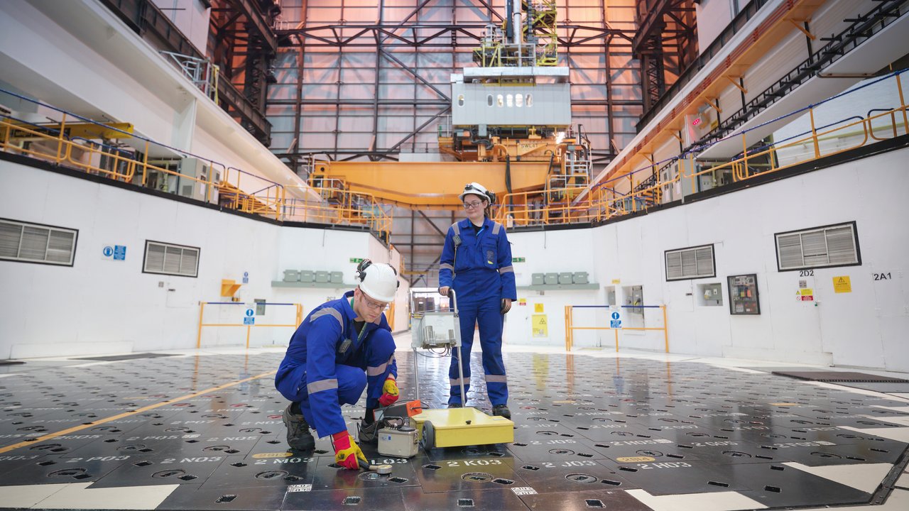 In a large industrial facility, two individuals in blue coveralls and safety gear are engaged in work on a metallic floor marked with various labels and symbols. One person kneels, inspecting a section of the floor, while the other stands nearby with a portable device. The expansive space features high ceilings and overhead equipment, emphasizing a technical environment.