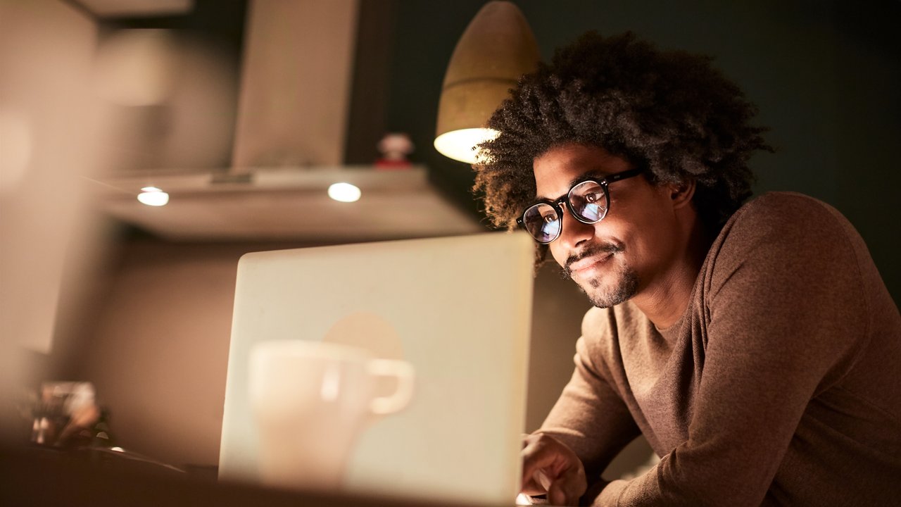 A person is seated at a table, focused on a laptop. The warm lighting from a pendant lamp creates a cozy atmosphere. A white coffee cup sits nearby, adding to the scene's comfort. The background features soft, blurred shapes, suggesting a modern interior space, enhancing the intimate, studious vibe.