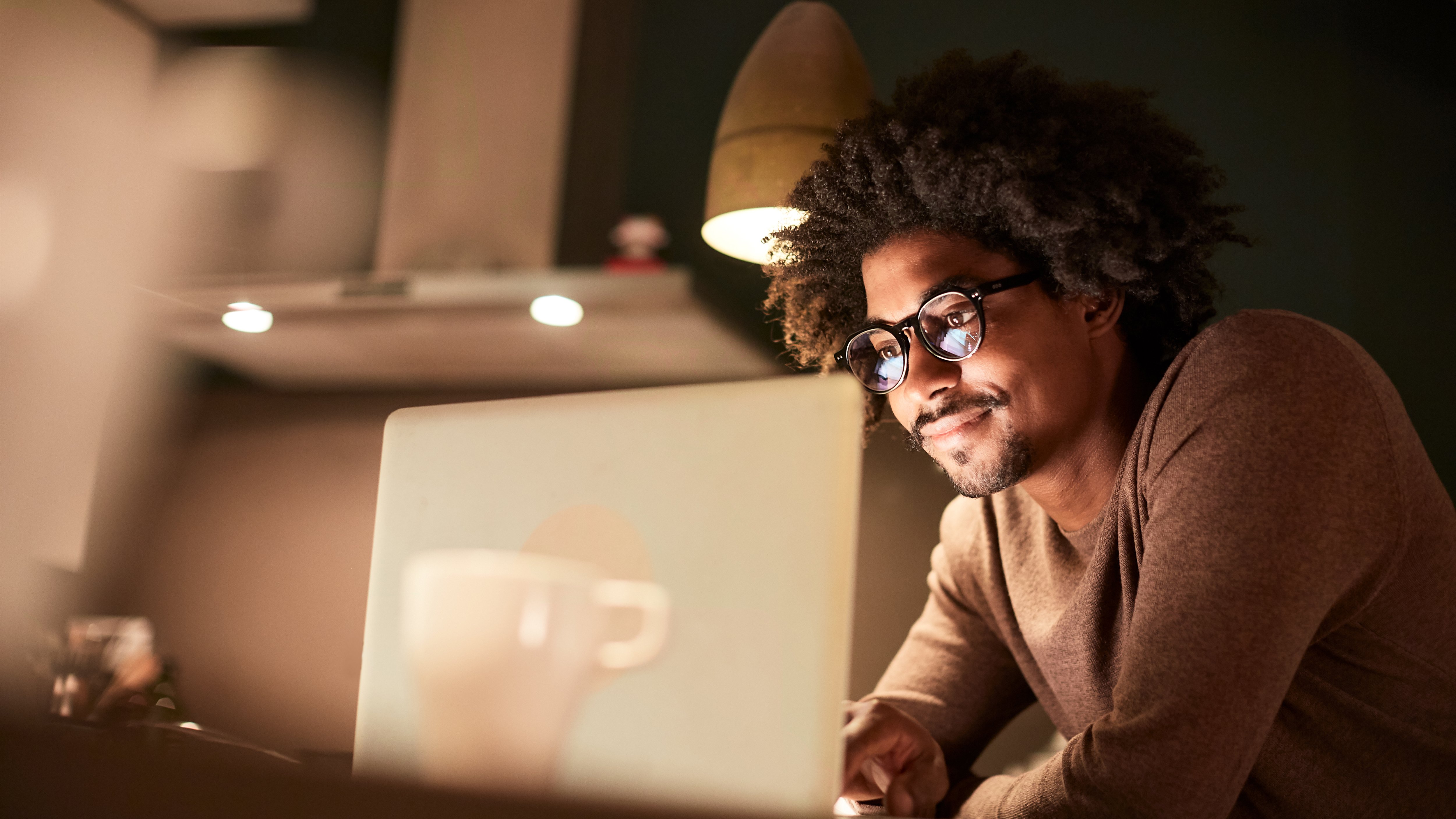 A person is seated at a table, focused on a laptop. The warm lighting from a pendant lamp creates a cozy atmosphere. A white coffee cup sits nearby, adding to the scene's comfort. The background features soft, blurred shapes, suggesting a modern interior space, enhancing the intimate, studious vibe.