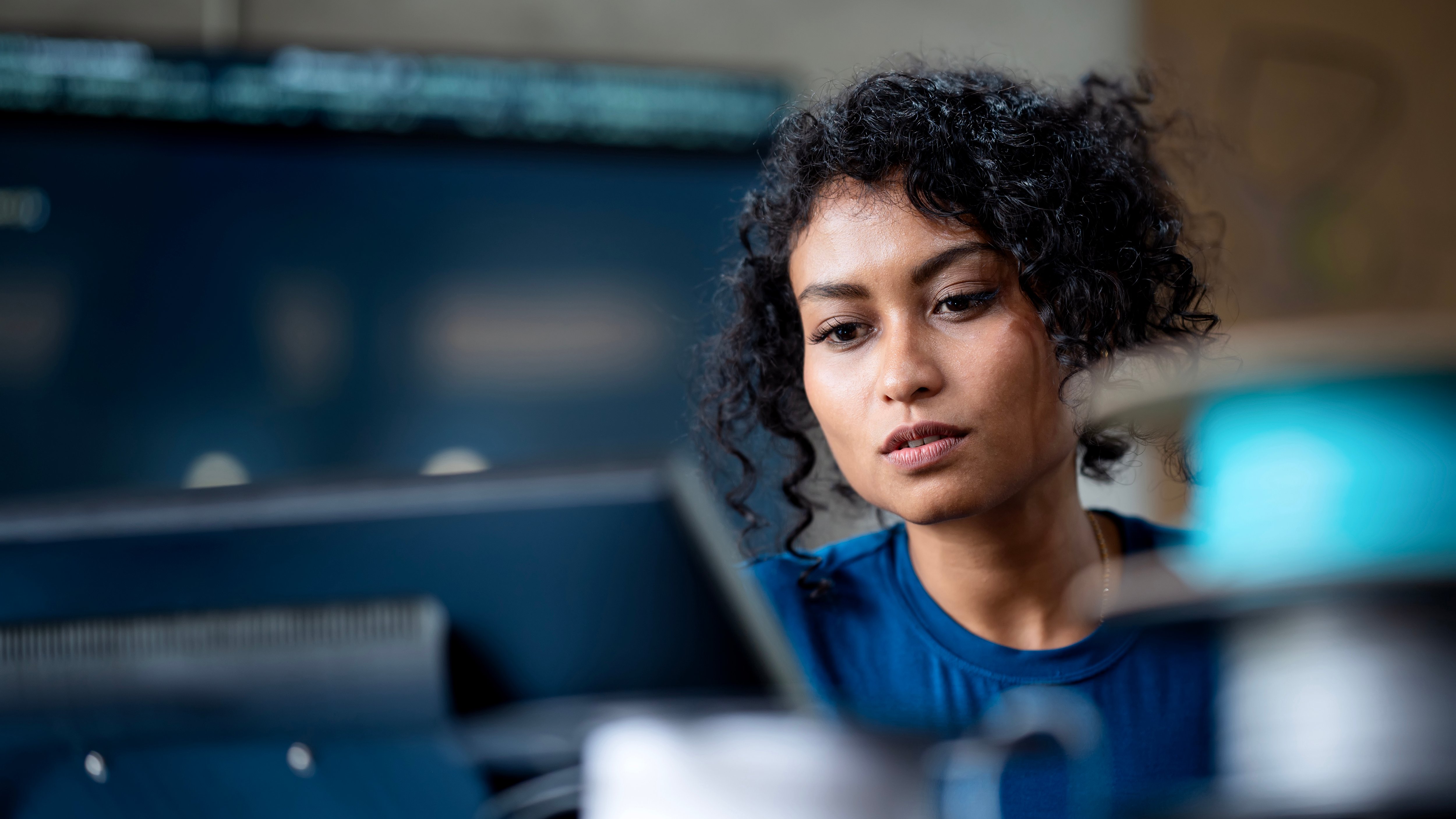A person with curly hair is seated at a desk, focused on a computer screen displaying code or data. They wear a blue shirt and are surrounded by various office equipment, including a monitor and cables. The background features a softly blurred workspace, enhancing the sense of concentration and productivity.