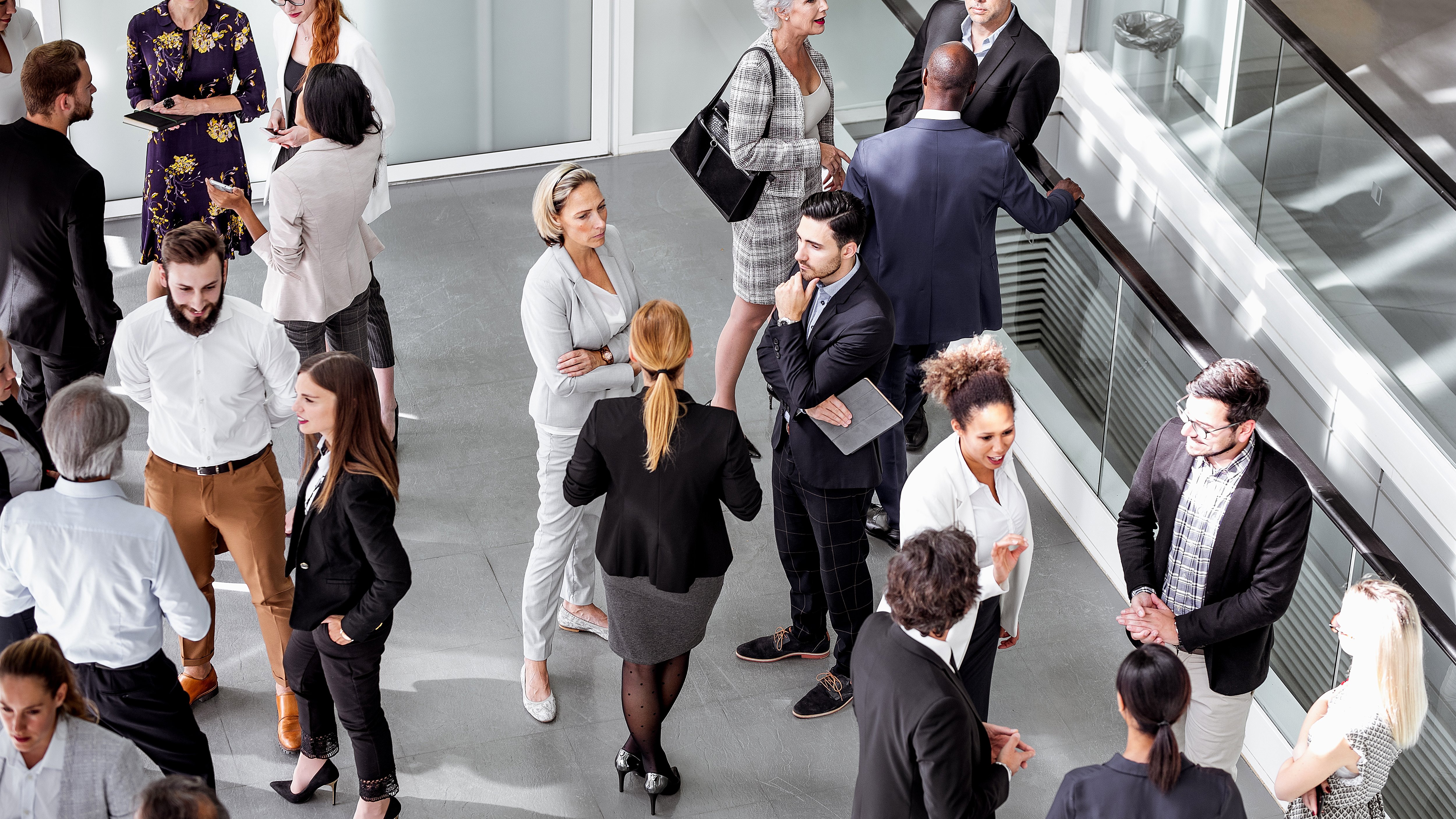A diverse group of individuals engages in conversation in a modern, well-lit space with large windows. Attire ranges from formal suits to business casual. Some hold notepads or devices, suggesting a professional context. The atmosphere is dynamic, with various clusters of people exchanging ideas and networking.