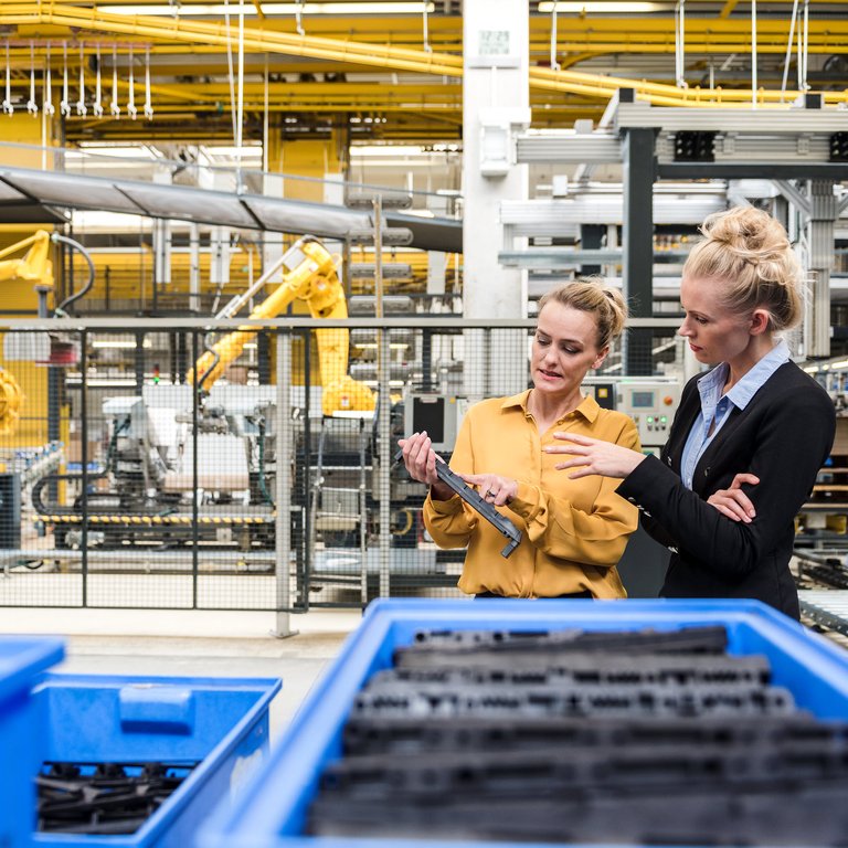 The Simple Six Two women discuss work while standing in a factory with robotic arms and blue bins in the background.