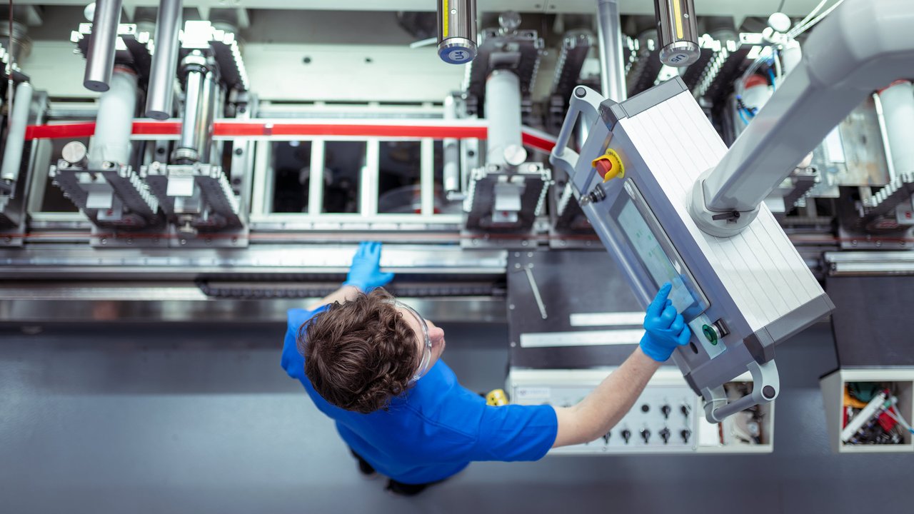 A worker in a blue shirt and gloves operates a control panel above machinery in a factory setting.
