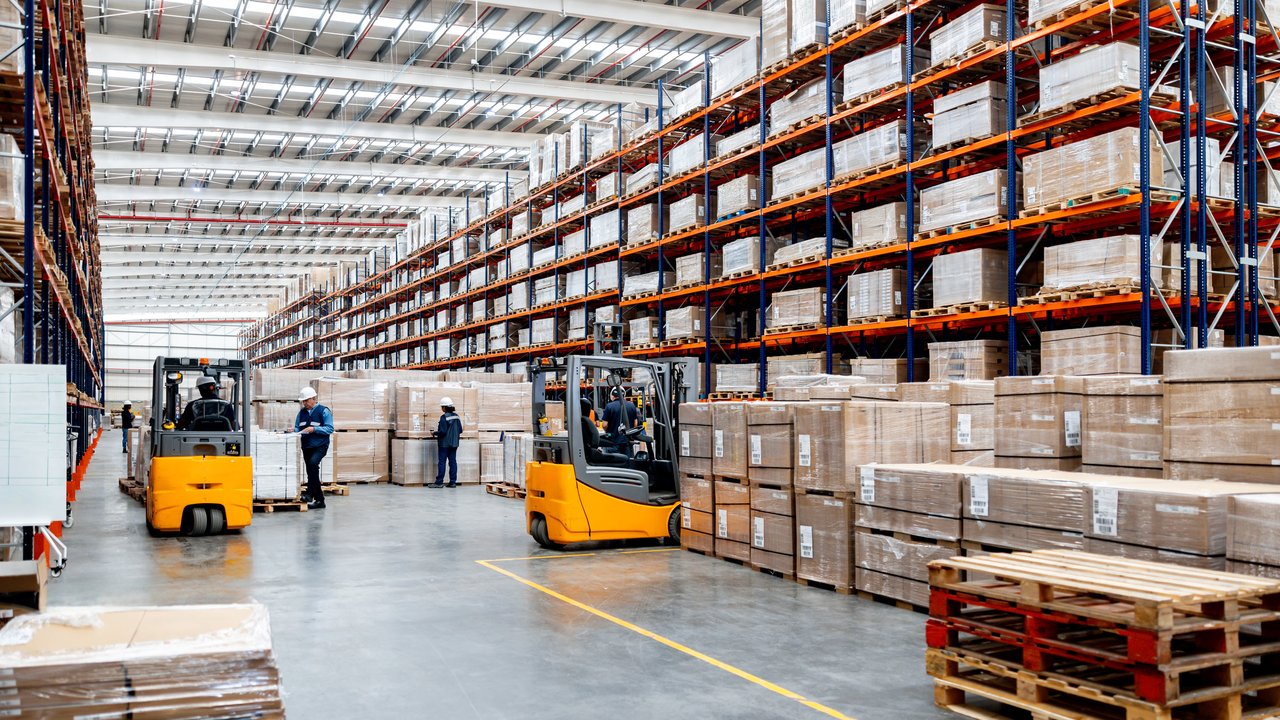 A warehouse with tall shelves stacked with boxes, two forklifts, and workers in safety gear. Brightly lit interior.