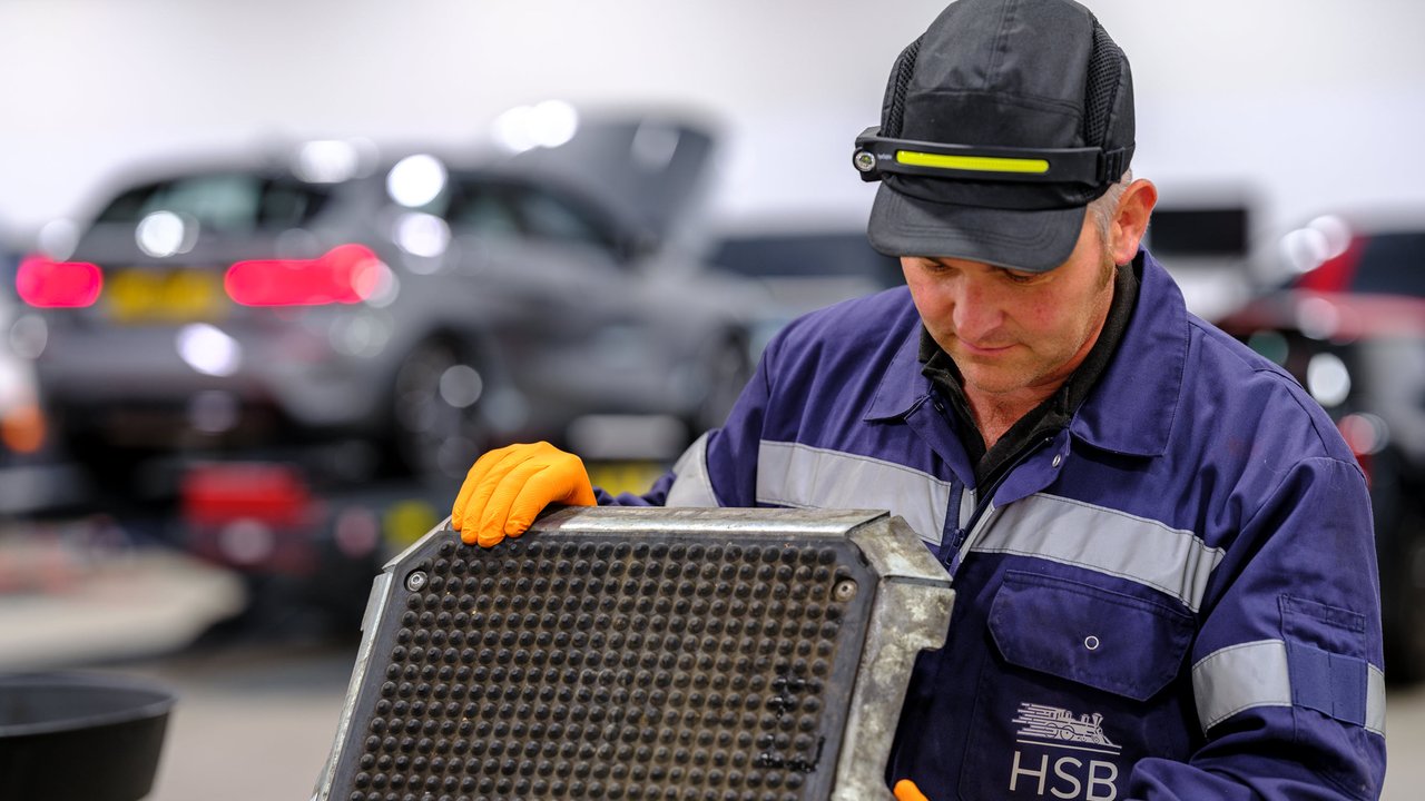 A HSB engineer surveyor in a blue uniform holds a large metal part in a garage with cars in the background.