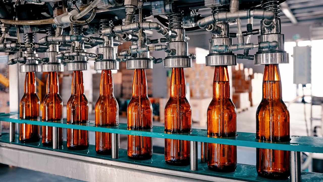 A row of amber glass bottles on a conveyor belt under a filling machine in a factory setting.