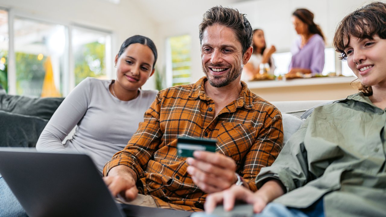 Three people sit on a sofa, one using a laptop and holding a credit card, with a kitchen visible in the background.