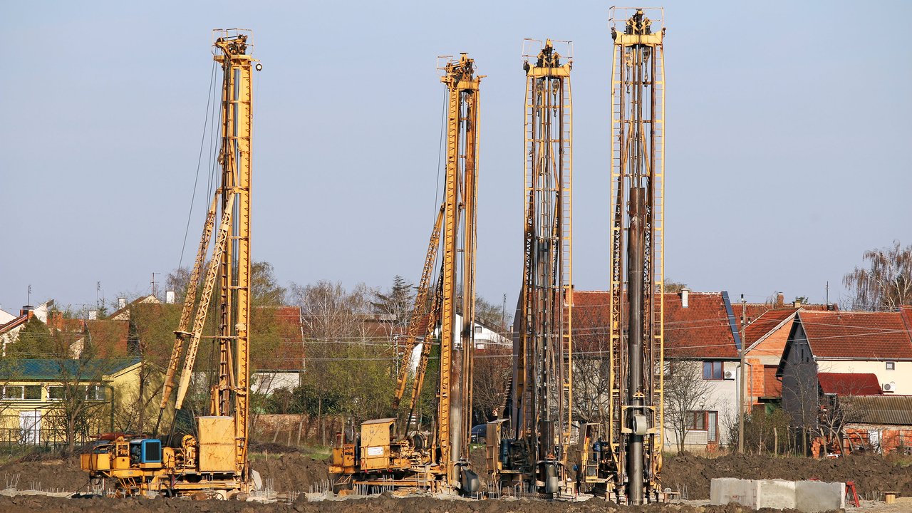 Four tall piling rigs are set up in a construction area near residential buildings.