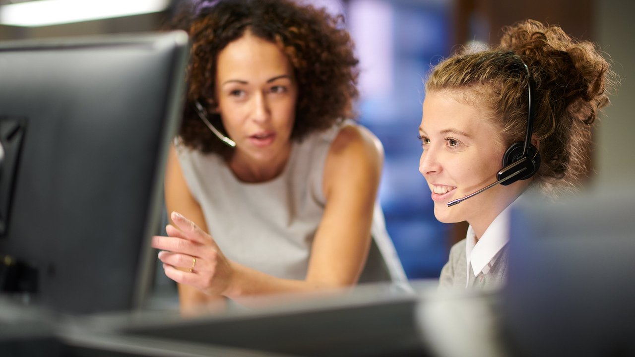 Claims Services Two women in a modern office setting, one pointing at a computer screen while the other listens with a headset.