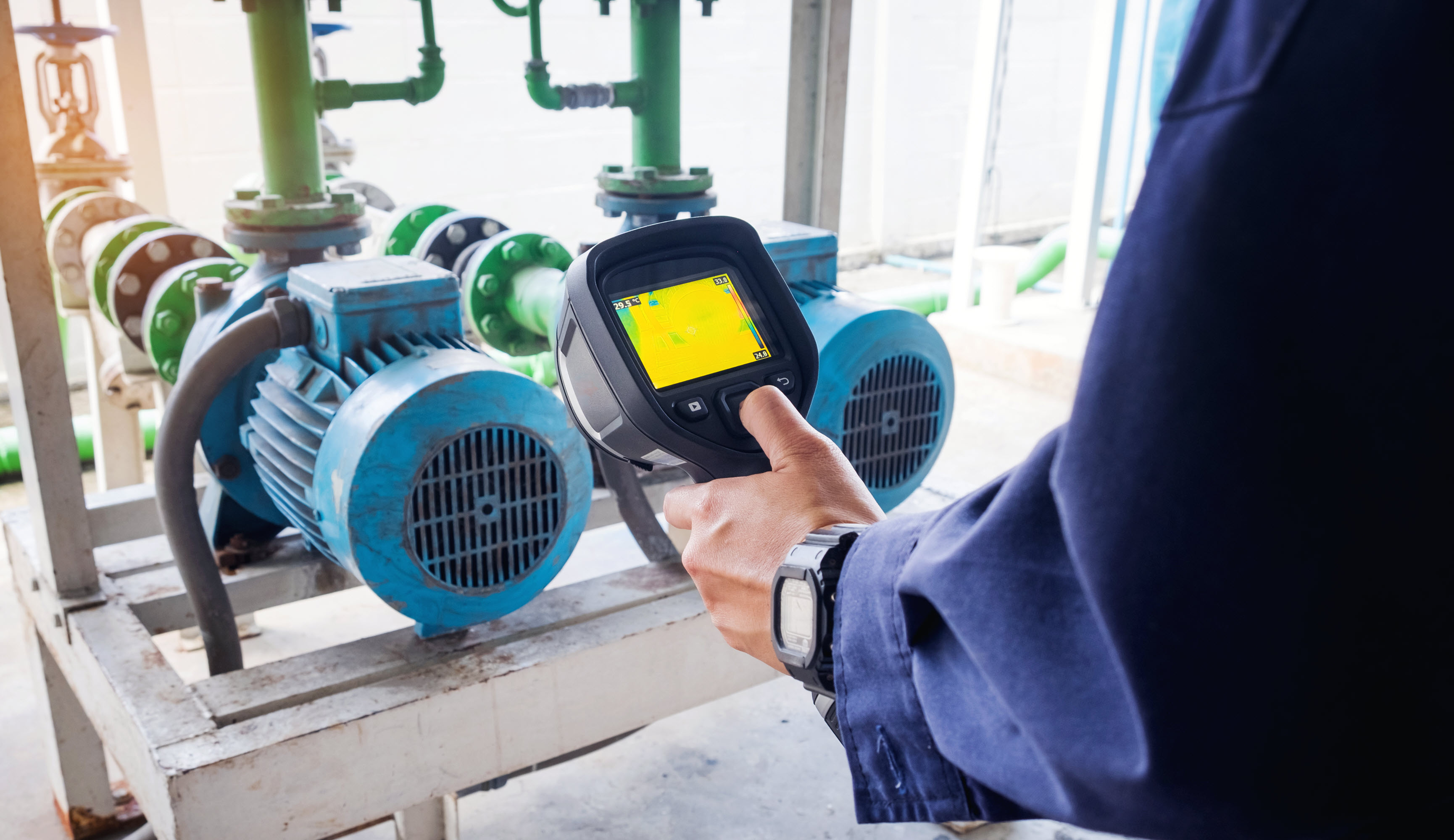 A person uses a thermal camera to inspect blue electric motors and pipes in an industrial setting.