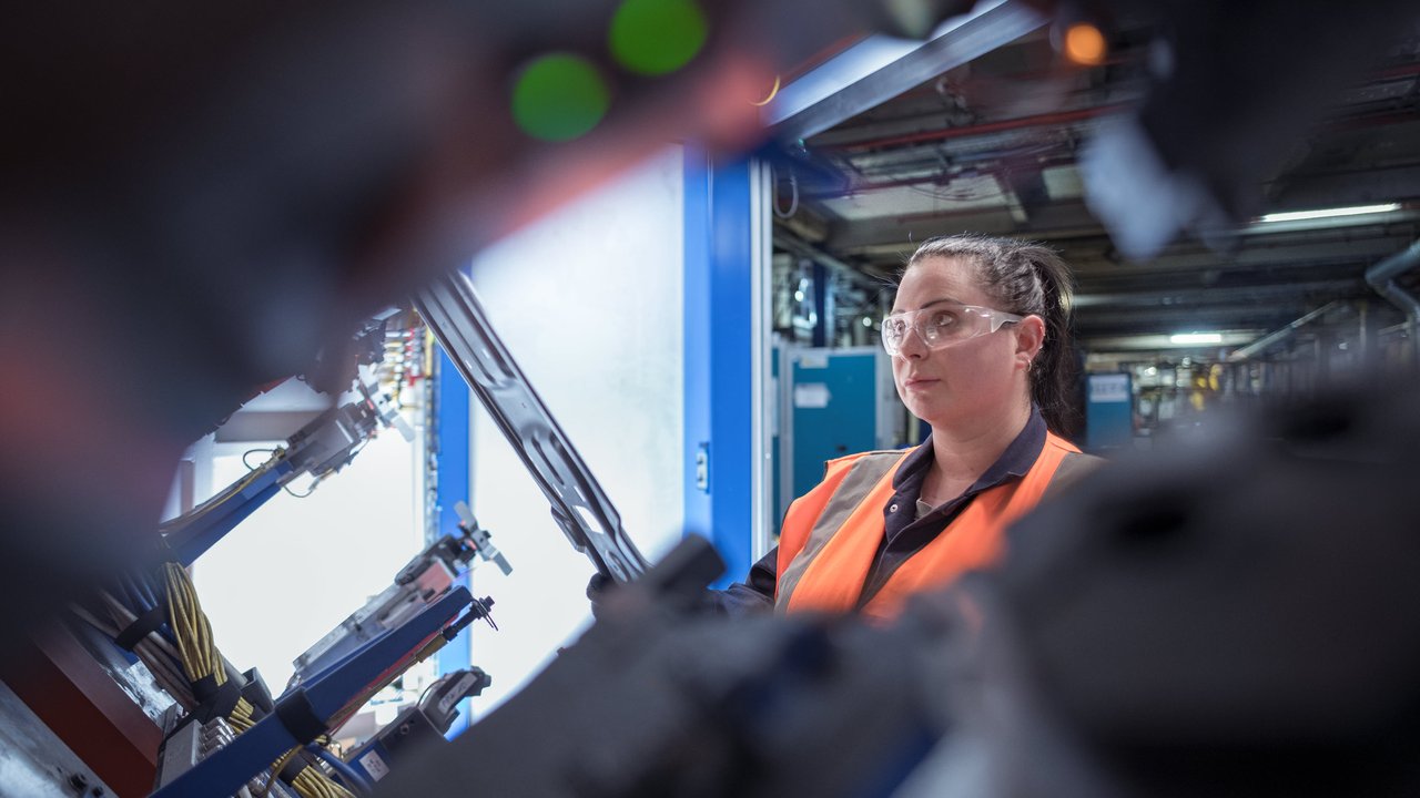 A person in an orange safety vest stands near machinery in a factory setting, with equipment and cables visible.