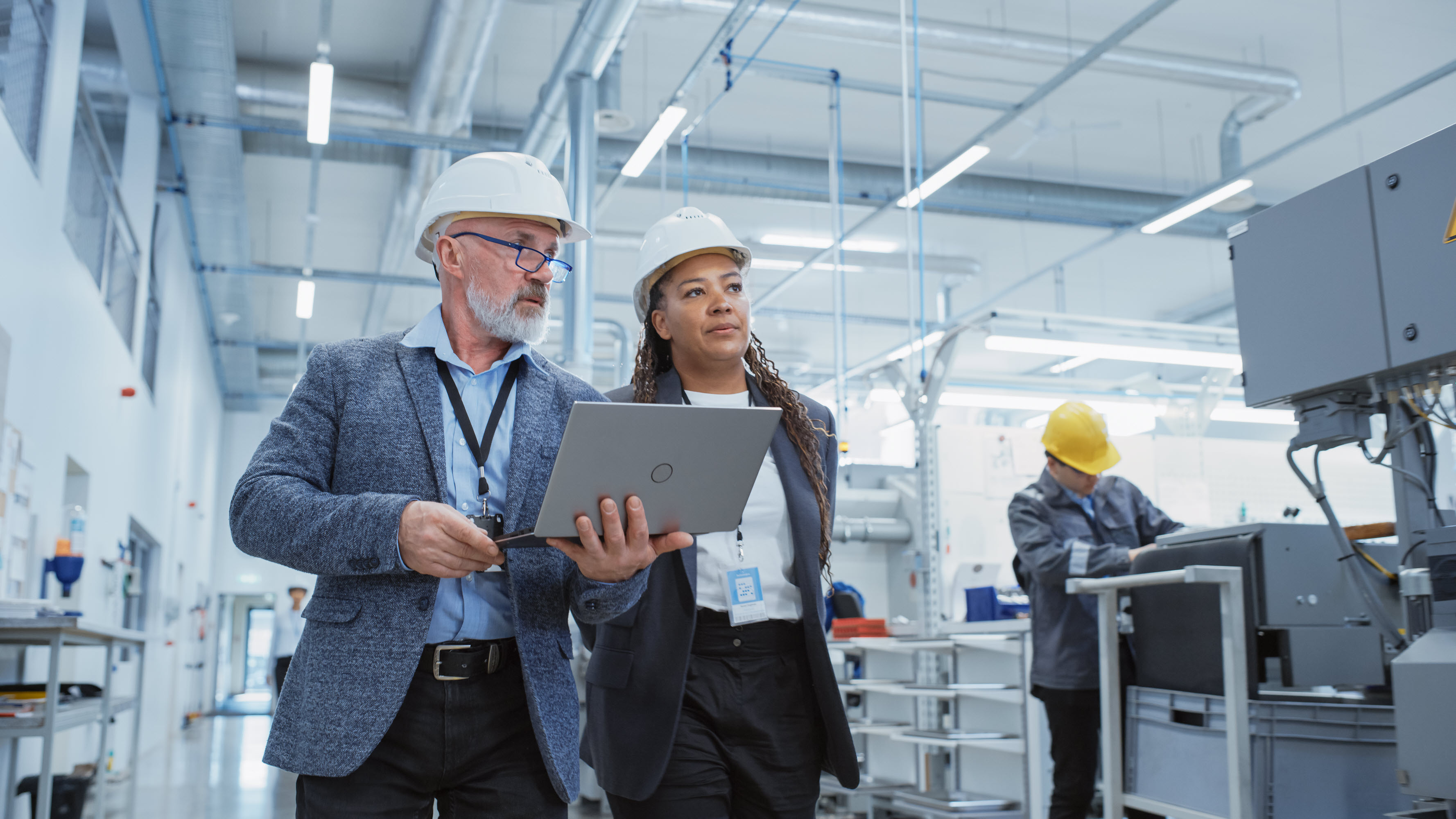 Two professionals in hard hats discuss over a laptop in a factory, while another worker operates machinery.