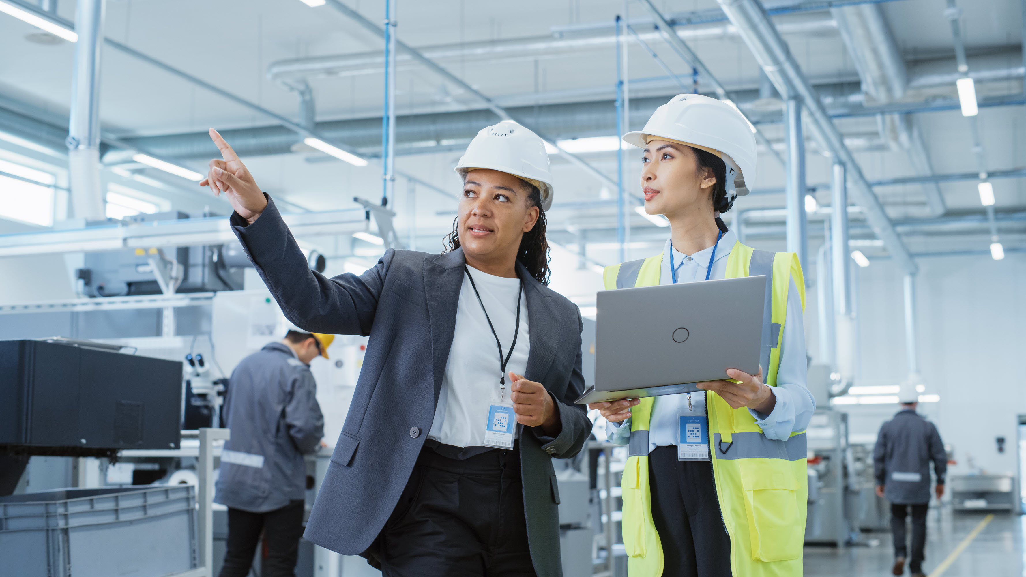 Two professionals in hard hats discuss while one points, with machinery and workers in the background.