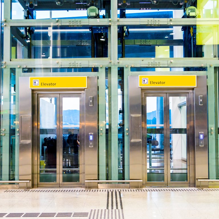 Two glass elevators with yellow signs labeled "Elevator" in a modern building lobby.