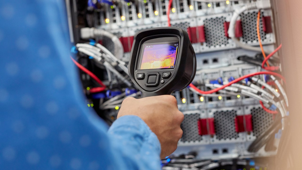 A person holds a thermal camera in front of server equipment with visible cables and lights.