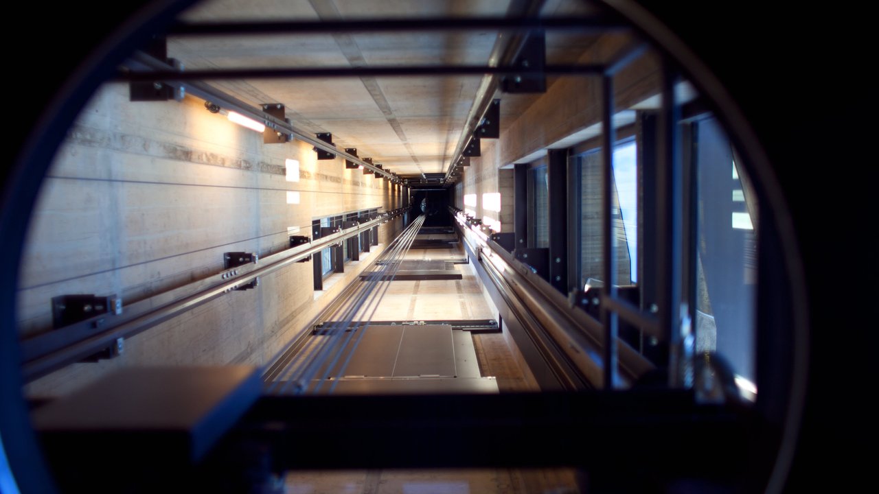 View of an elevator shaft with metal tracks and concrete walls, illuminated by overhead lights.