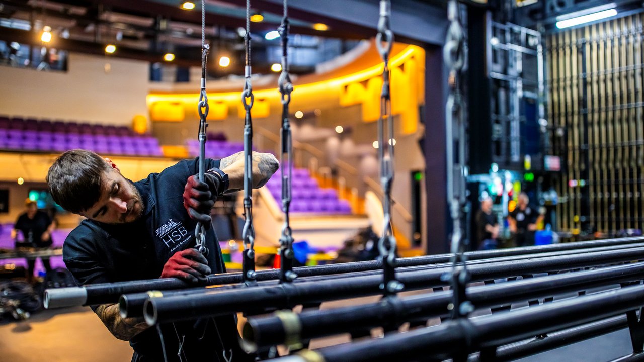 A technician adjusts rigging equipment on stage in a theater with purple seating in the background.