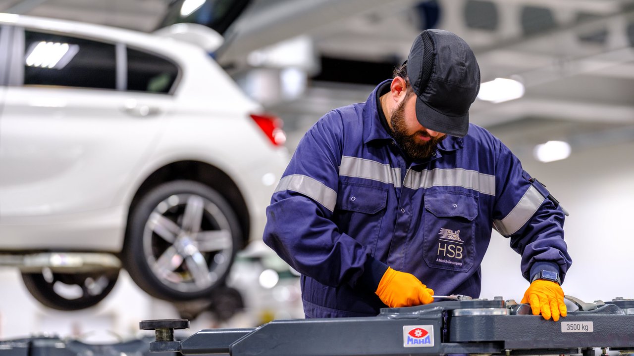 A mechanic in a blue uniform works on equipment in a car service center, with a white car elevated in the background.