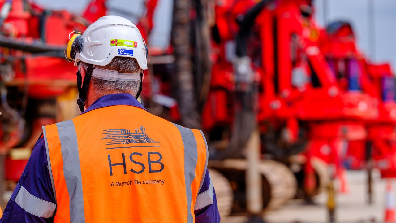 A worker in an orange safety vest and helmet stands in front of large red machinery on a construction site.