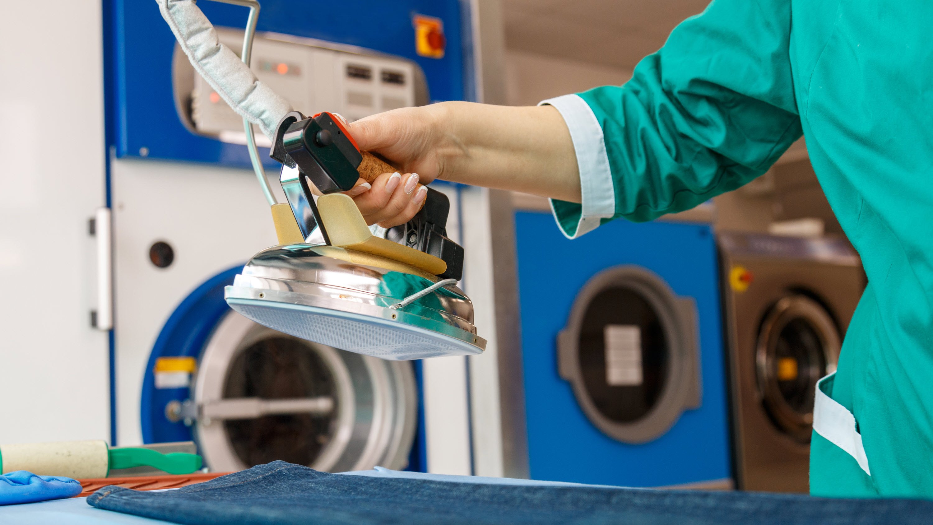 A person in a green uniform uses a steam iron on a pair of jeans in a laundry setting.