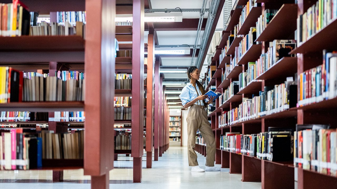 A person stands in a library aisle, holding a book, surrounded by tall shelves filled with books.