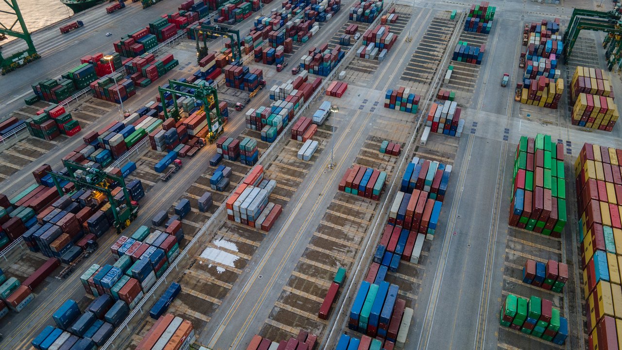 Aerial view of a shipping port with numerous stacked cargo containers and cranes, alongside empty parking spaces.